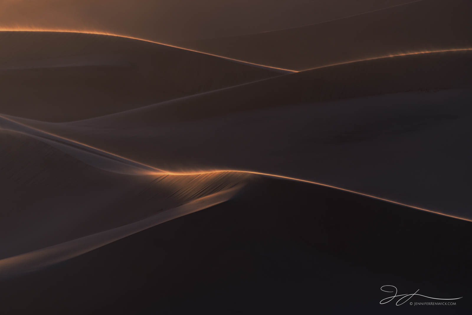 Light highlights the ridges of sand dunes during a windy sunset.