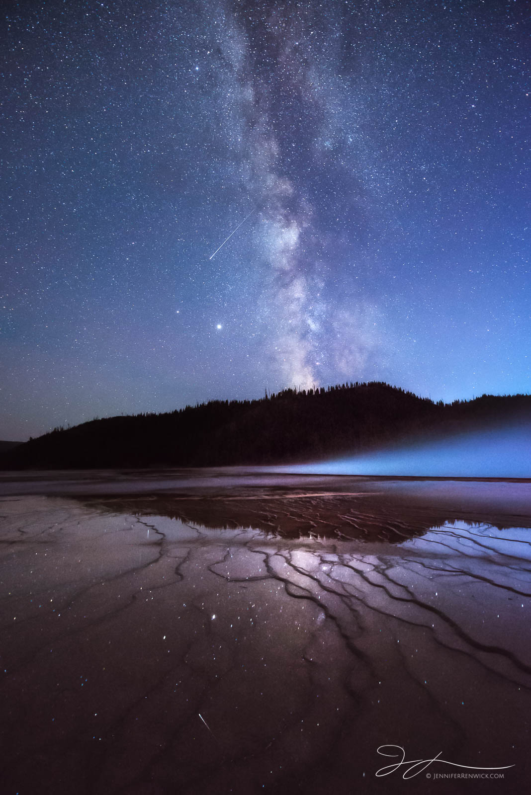 The terraces around Grand Prismatic Spring reflect the night sky, while a meteor passes through the milky way.